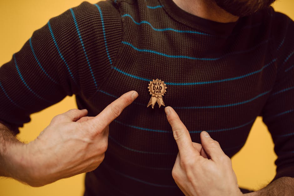 A man proudly highlighting his 'Best Dad Ever' badge on a striped sweater against a yellow background.