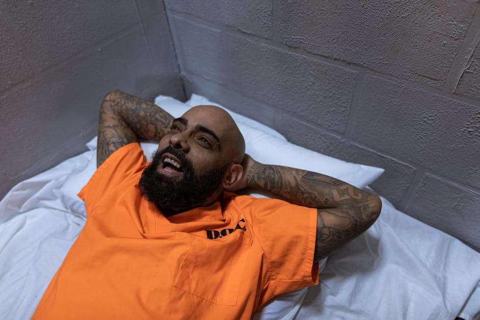Tattooed man in orange jumpsuit smiling while lying in a prison cell.