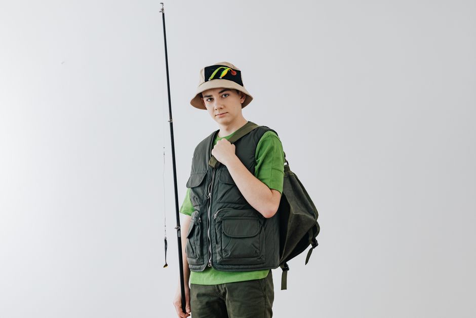 Teenager in green gear with fishing rod and hat against a white background.
