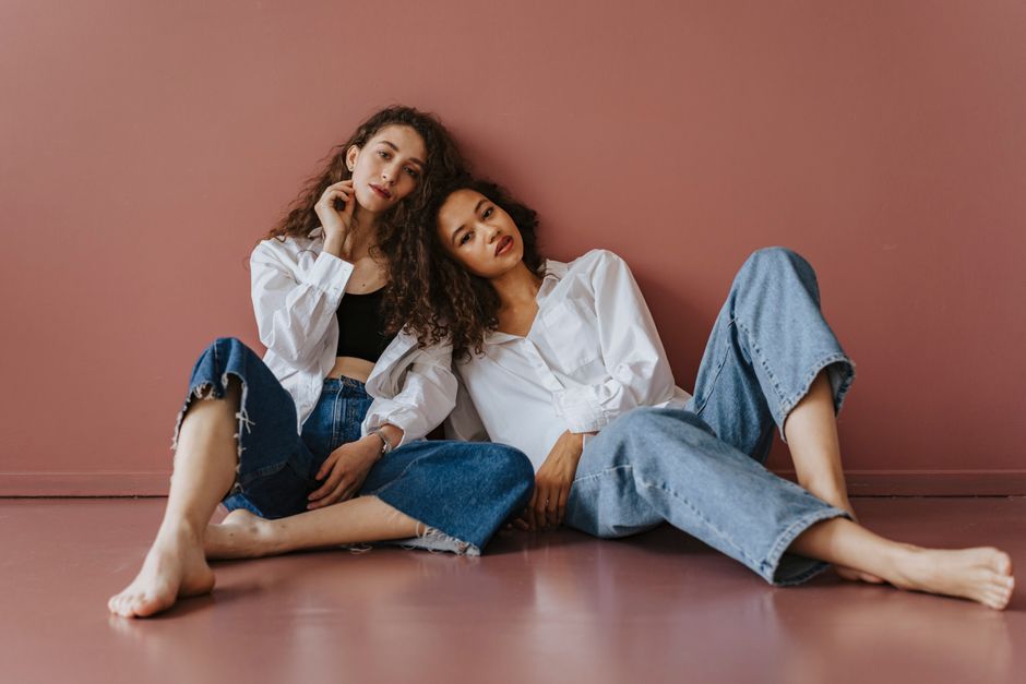 Two women sitting against a pink wall, dressed in denim and white shirts, conveying casual elegance.