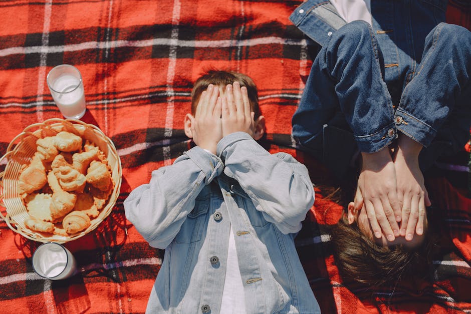 From above of boy and girl in jean jackets lying on plaid blanket with faces closed hands near glasses of milk and pastry in wicker basket