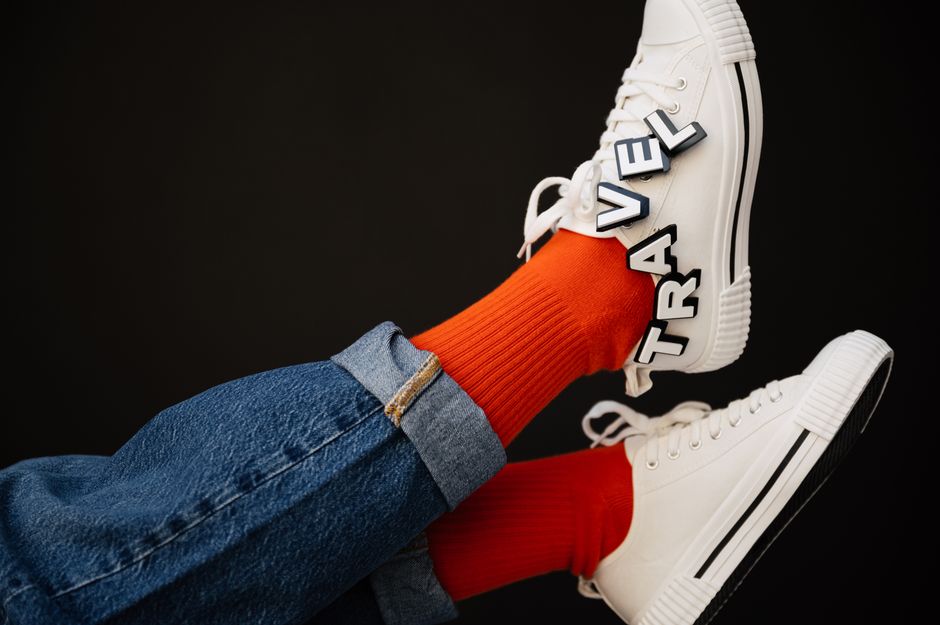 Close-up of white sneakers with 'TRAVEL' letters, paired with orange socks and denim jeans.