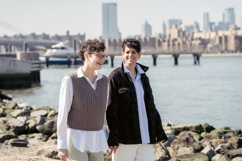 Delighted young homosexual ladies with short dark hair in stylish clothes laughing while holding hands and walking near city port on stony river embankment