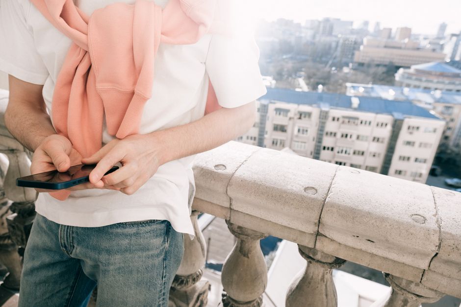 A young man stands on a balcony using his smartphone, overlooking the city.