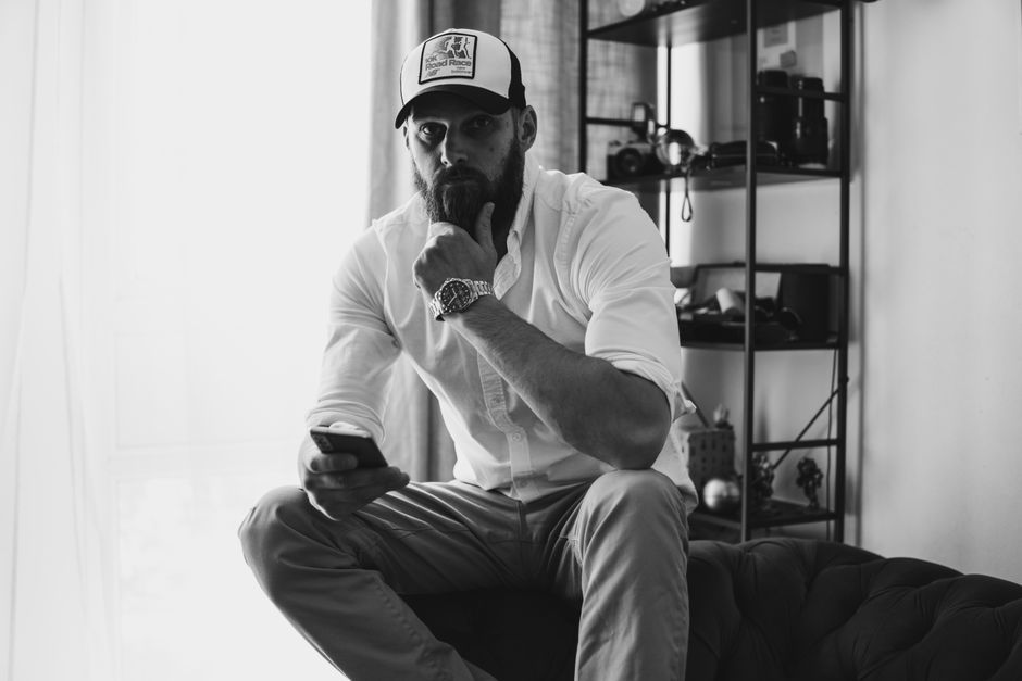 A thoughtful man sitting indoors, holding a phone, captured in a stylish black and white portrait.