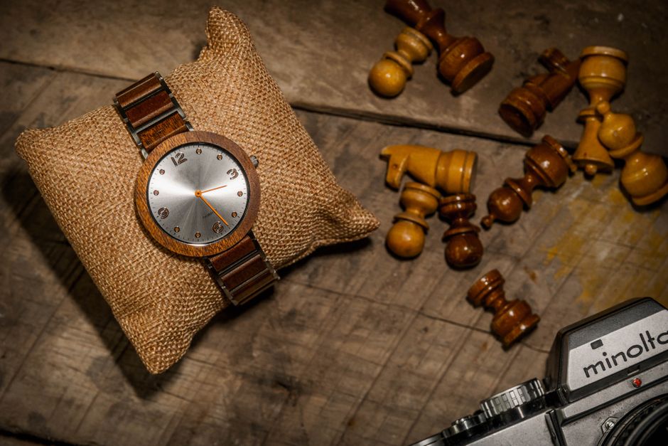 Vintage wooden wristwatch on burlap pillow beside scattered chess pieces and Minolta camera on rustic table.