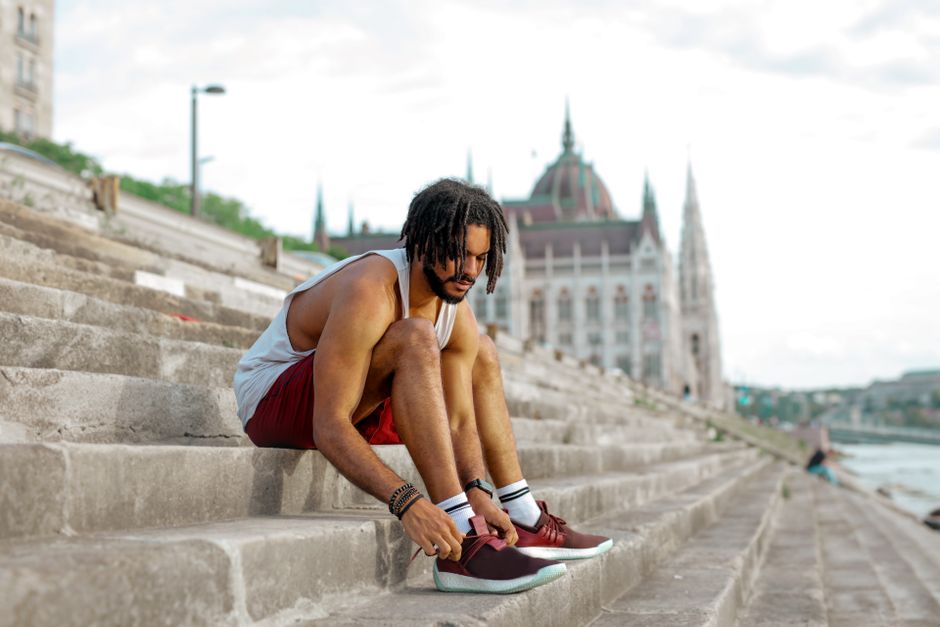 Man with dreadlocks tying shoes on concrete stairs near a recognizable European landmark.