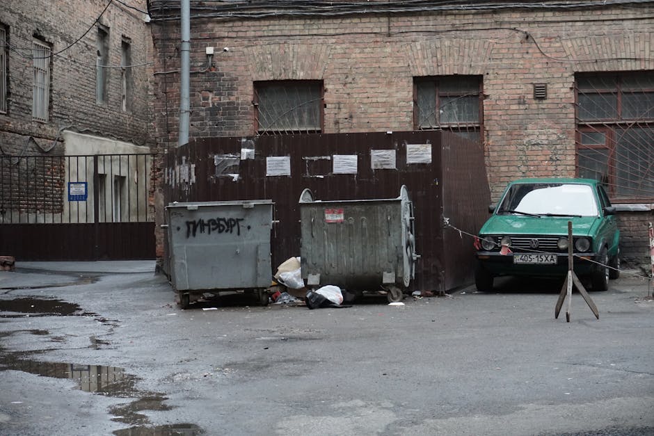 A gritty urban alley featuring garbage bins, scattered litter, and an old parked car.