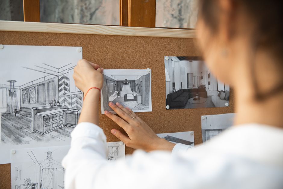 Person arranging interior design sketches on a corkboard indoors.