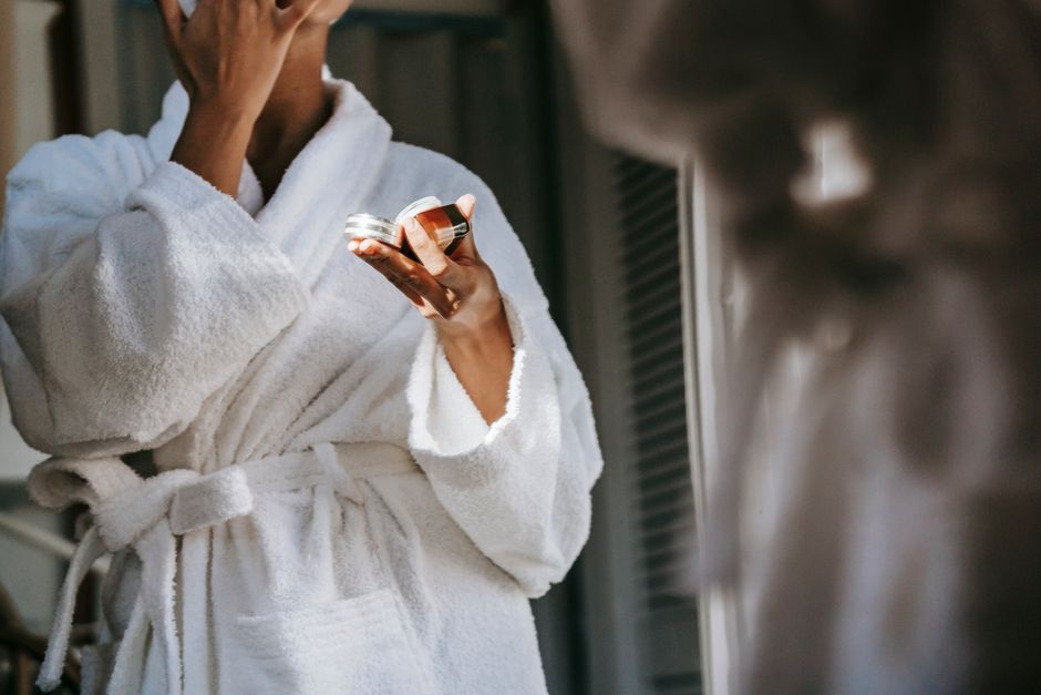 Crop anonymous ethnic female in soft bathrobe applying nourishing cream on face while refl