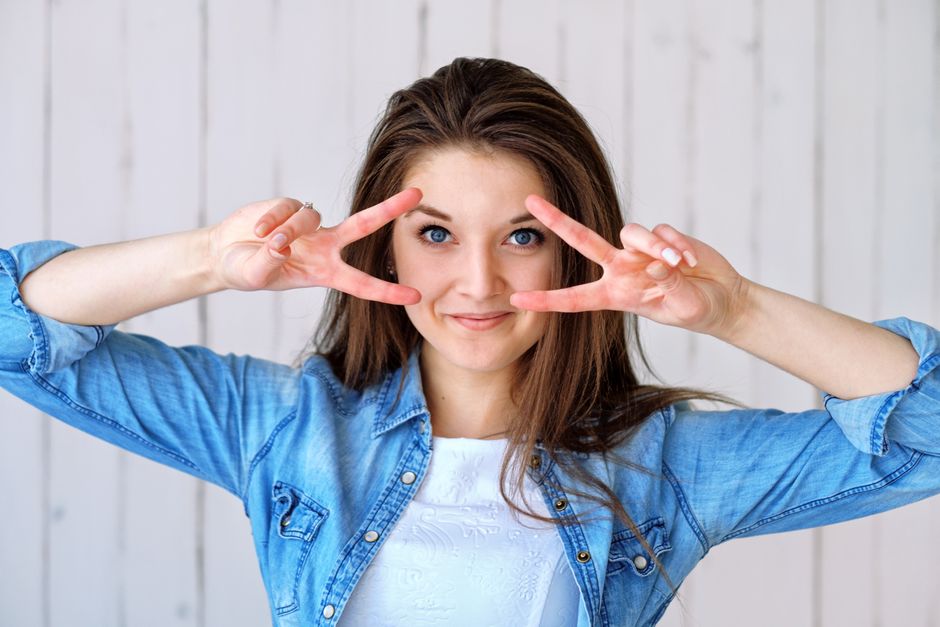 Cheerful woman in denim making a peace sign indoors. Captured in a bright and casual setting.