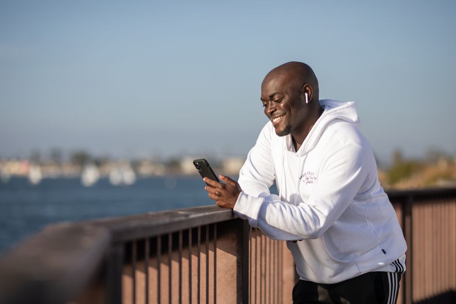 A man in a white hoodie smiles while using a smartphone outdoors by the railing.