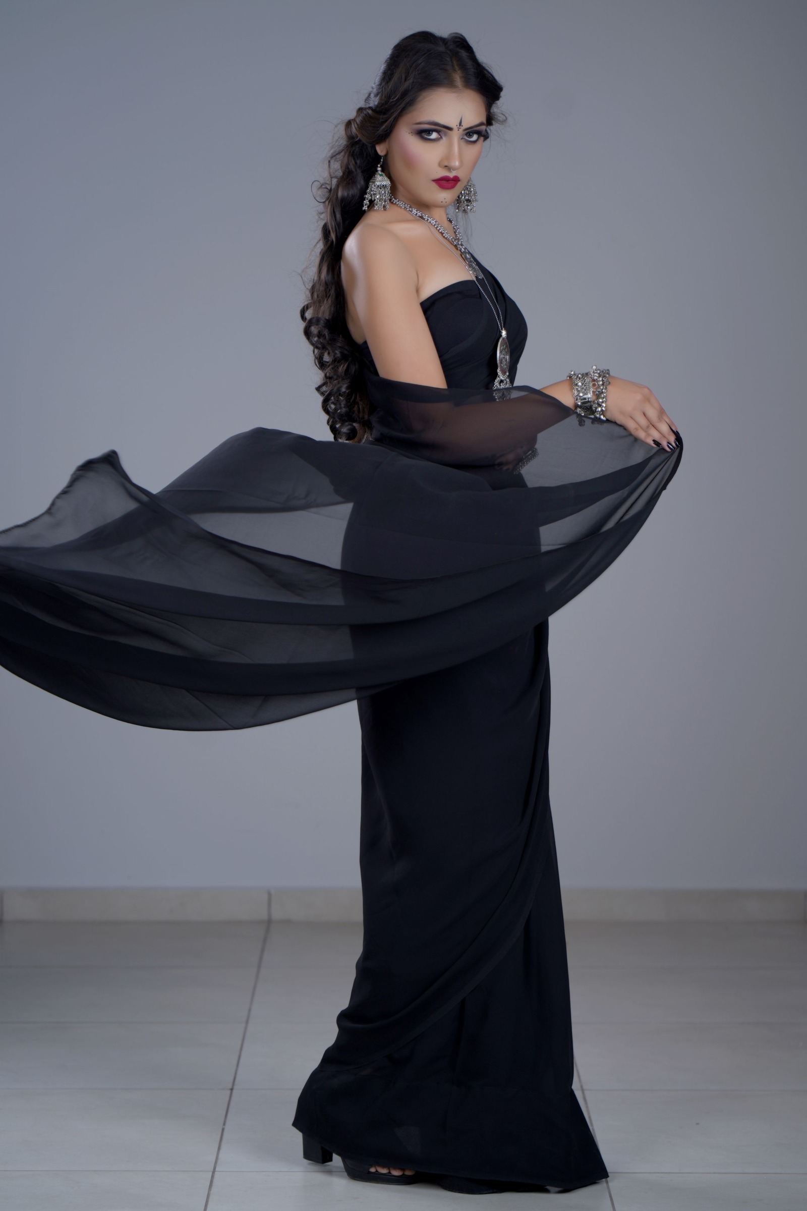 Full-length studio view of a woman in a black evening gown with a long sheer panel held out to the side against gray.