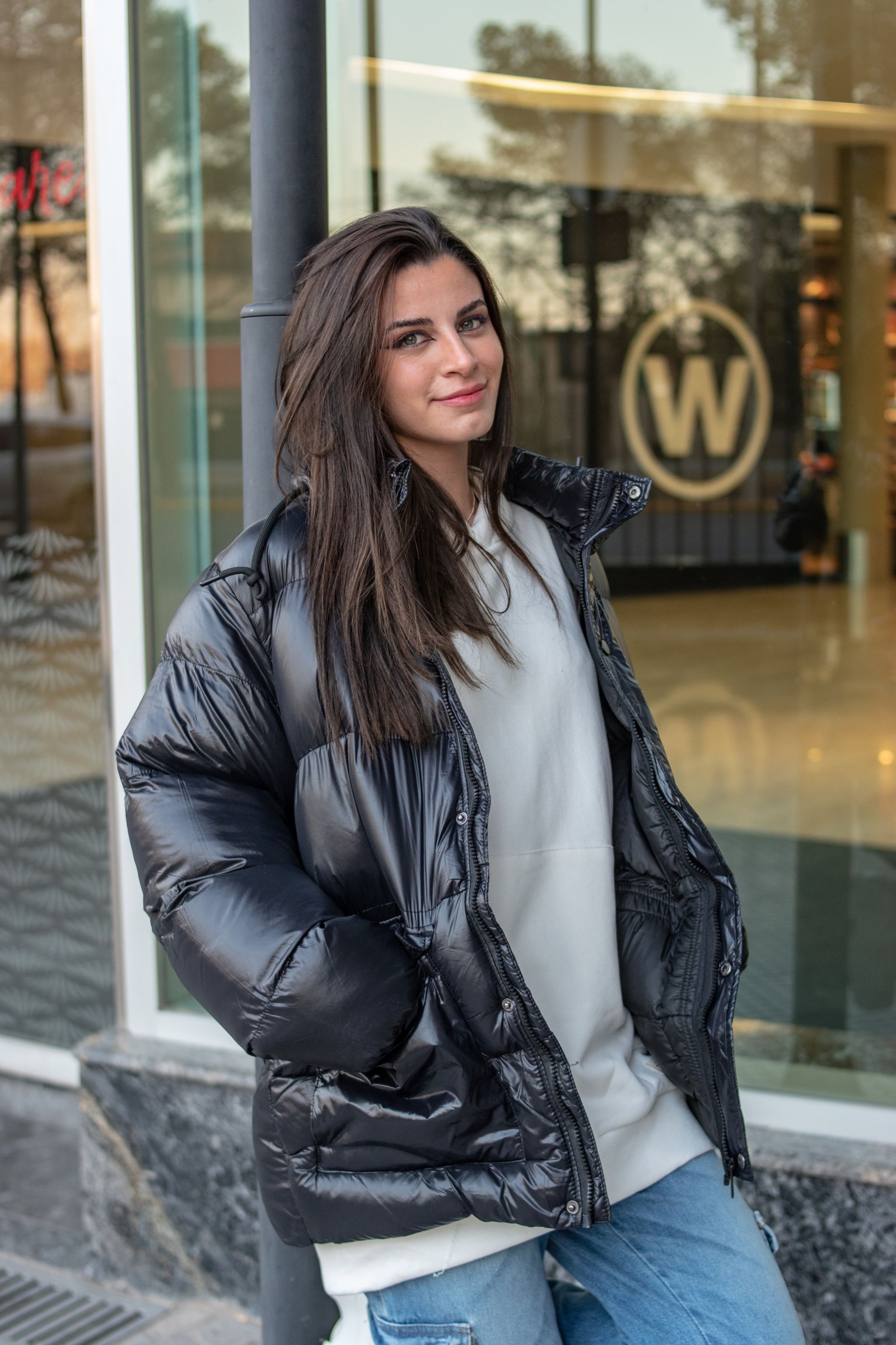Woman in an open black puffer jacket over a gray sweatshirt and jeans on a city sidewalk.