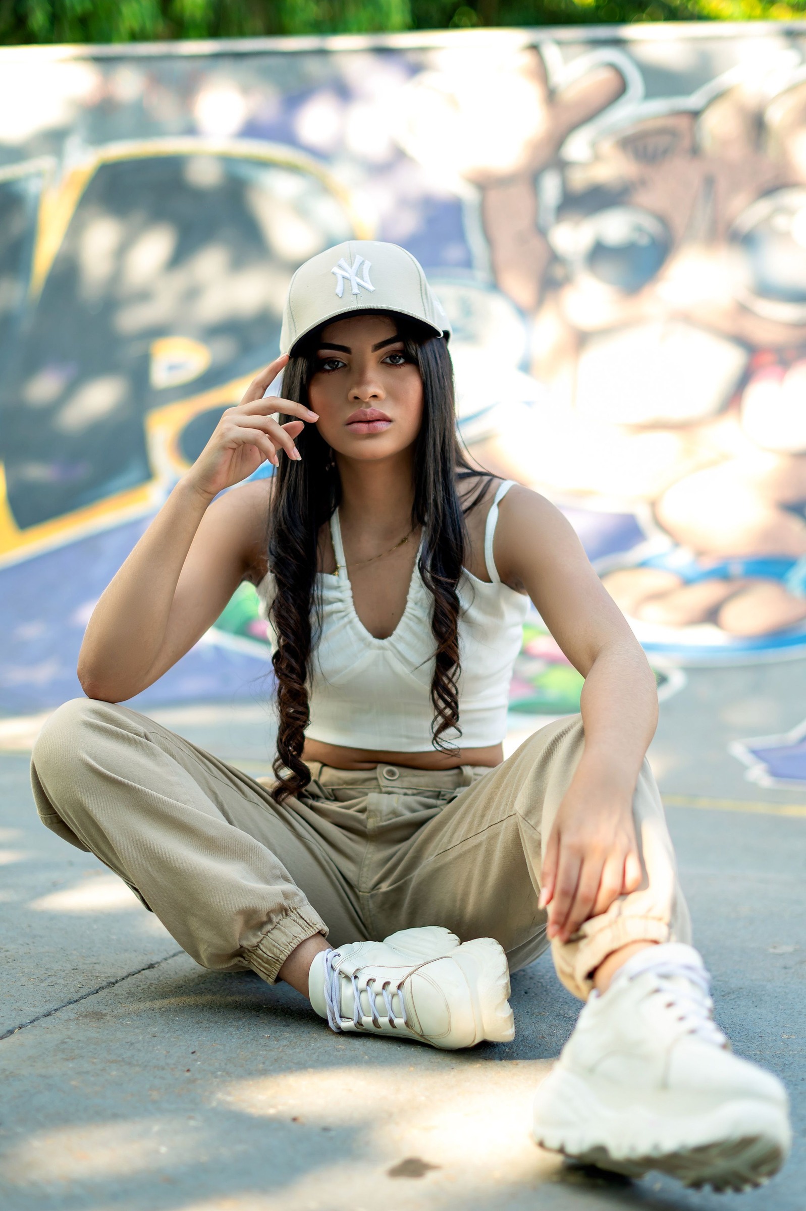 Woman in a gray embroidered baseball cap, white sleeveless top and loose khaki trousers seated on concrete by a graffiti wall. Illustrative cap, not Pokemon merchandise.