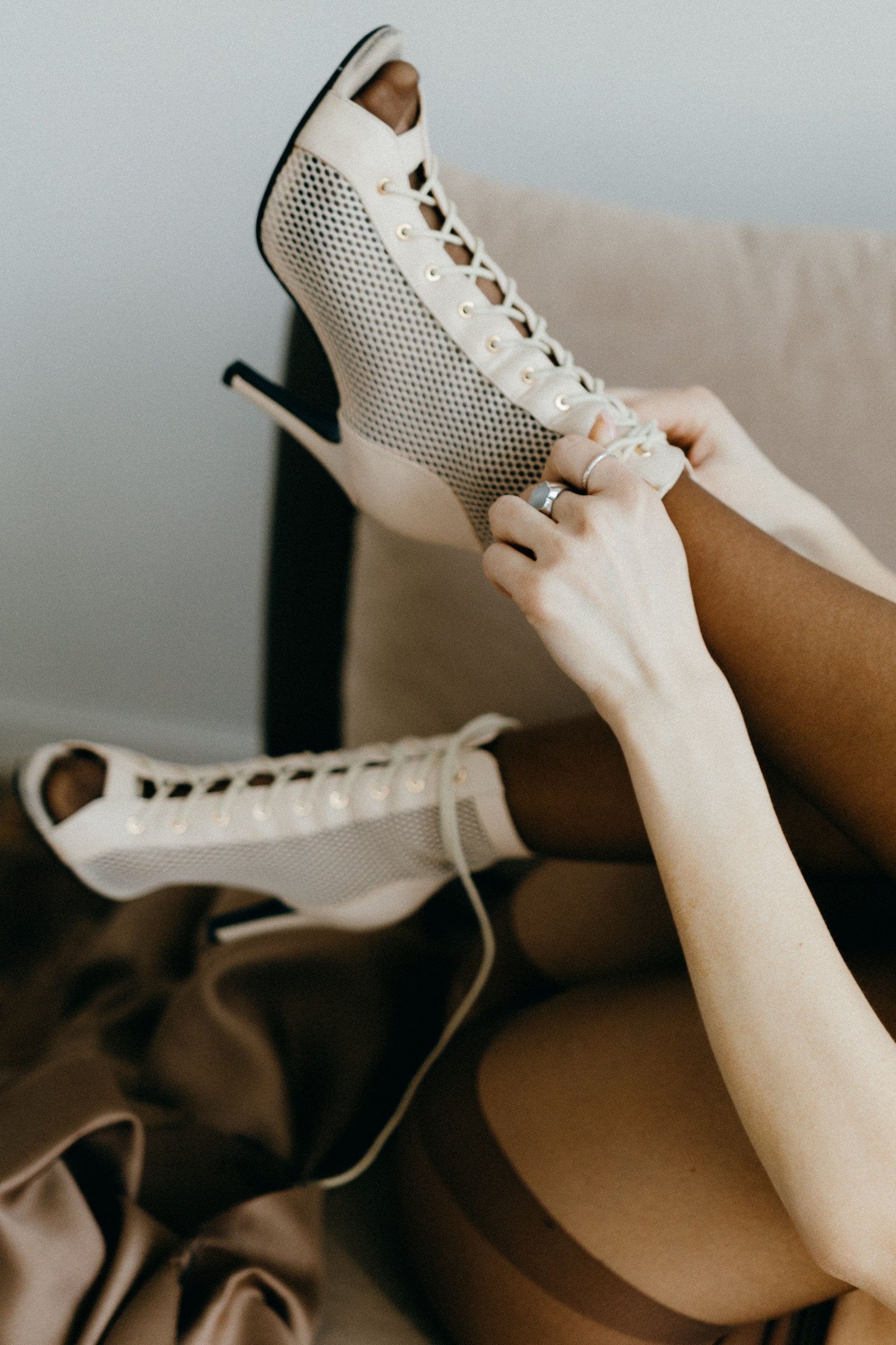 Close-up of hands fastening pale beige mesh peep-toe lace-up booties with stiletto heels; footwear and lower legs only.