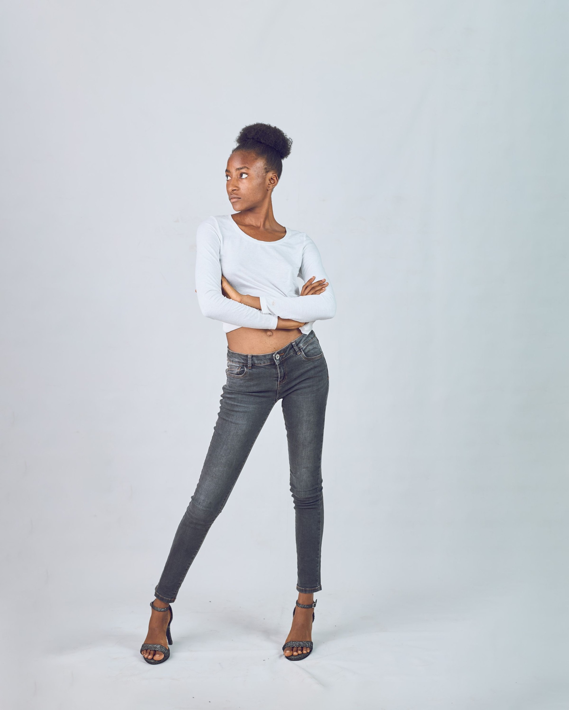 Full-length studio view of a woman wearing dark skinny jeans, a white cropped long-sleeve top and heeled sandals.