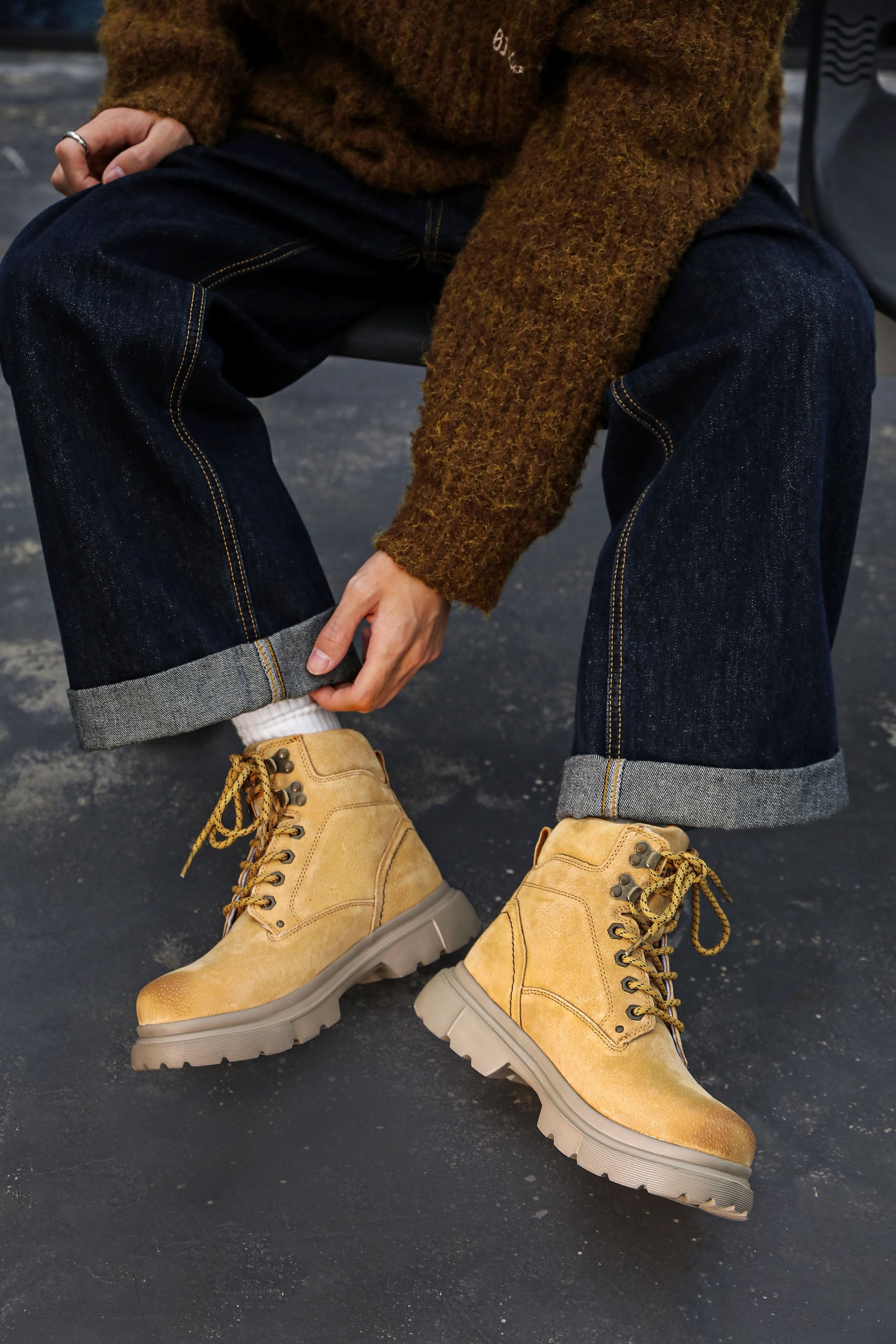 Cropped seated view of loose dark jeans with broad cuffs above tan lace-up boots on concrete.