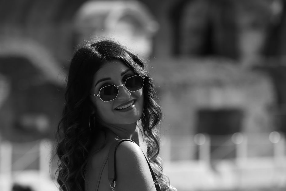 Black and white image of a woman smiling while wearing sunglasses, captured outdoors.