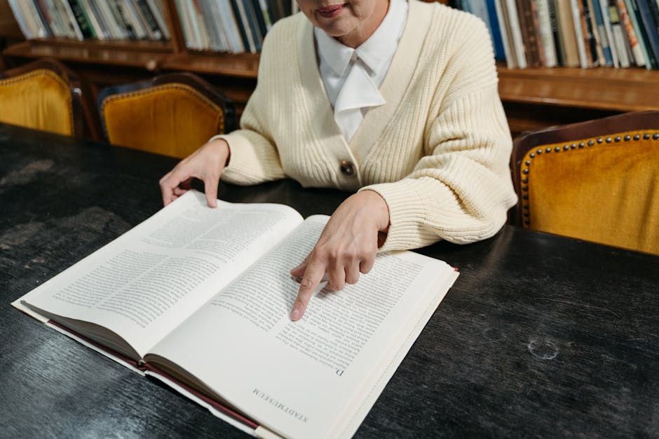 Woman pointing to text in an open book at a library's dark wooden table.