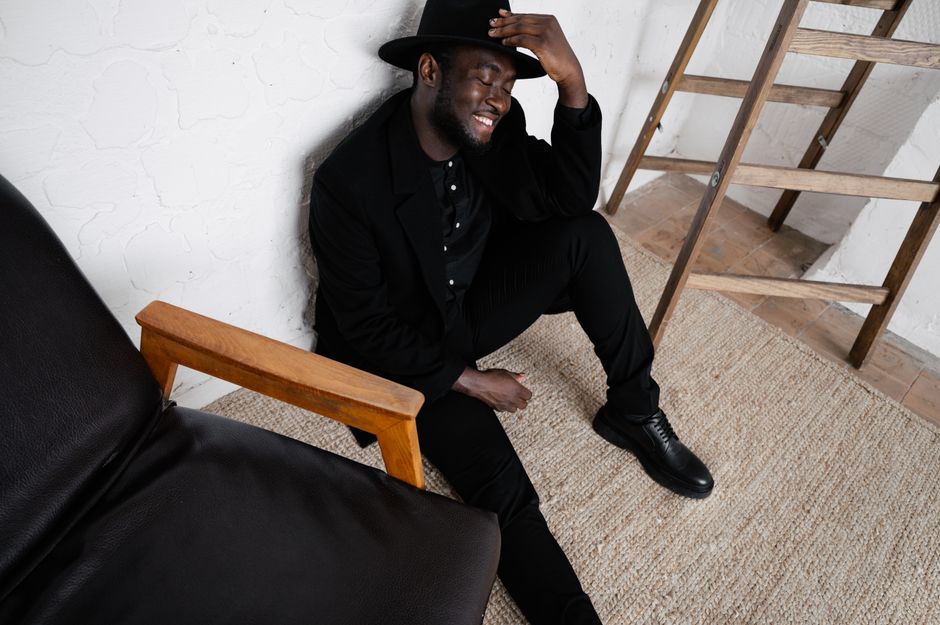 From above of trendy African American male in black outfit sitting on floor on beige rug near armchair while smiling and touching hat with fingers with eyes closed