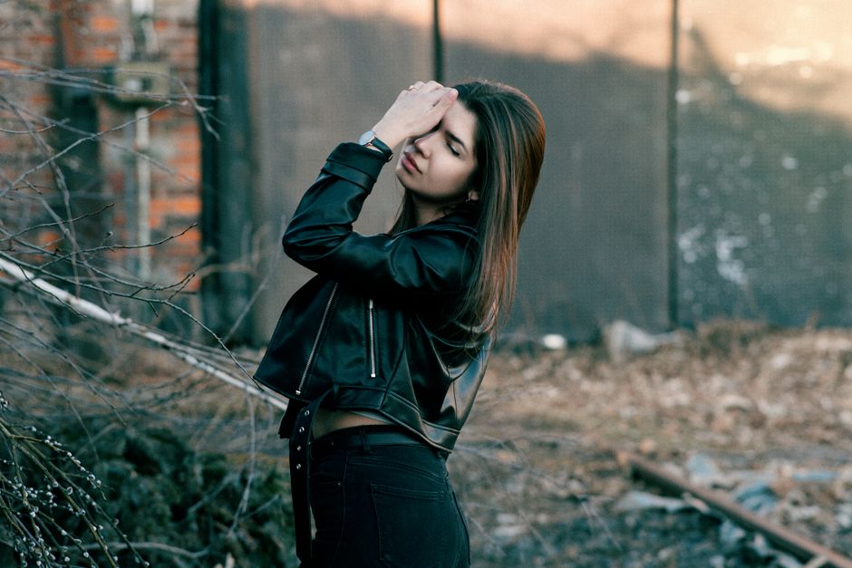 A young woman wearing a leather jacket poses with eyes closed in an outdoor setting.