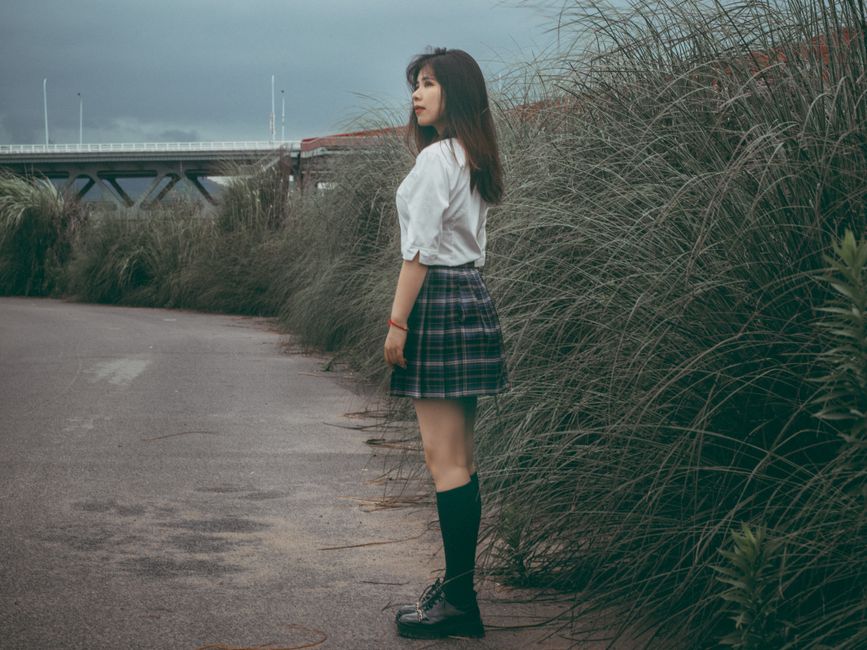 A young woman standing on a walkway lined with tall grass, under an overcast sky.