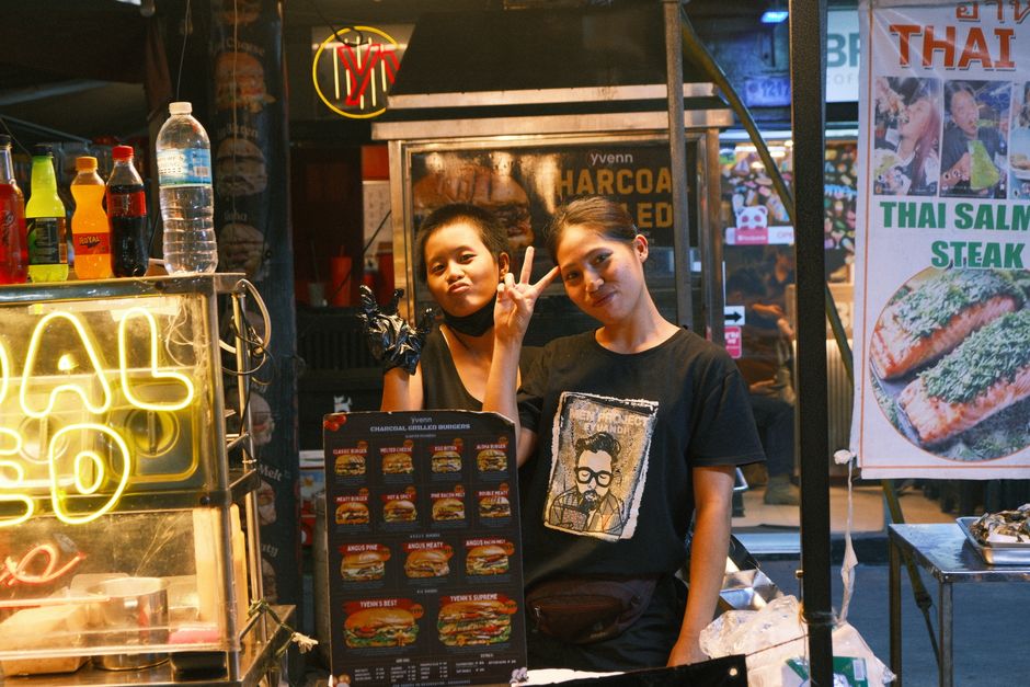 Two women pose at a lively Thai street food stall with neon signs and delicious menu options.