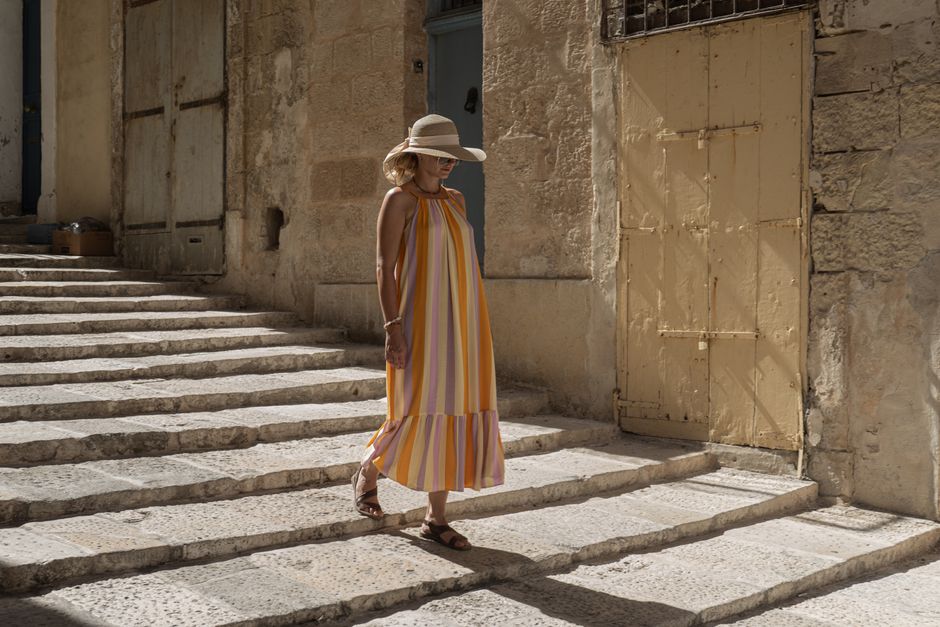 A woman in a sun hat and striped dress walks down stone steps in Valletta, Malta.