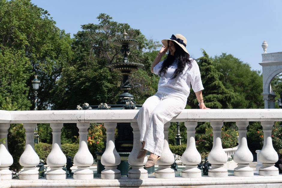 Woman sitting on marble railing in Baku park, enjoying a sunny day. Lush greenery and elegant fountain in background.