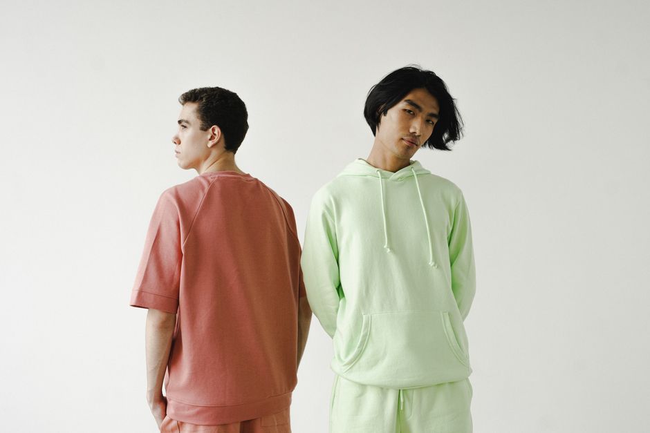 Two men standing back-to-back in pastel outfits against a white wall, showcasing minimalist fashion.