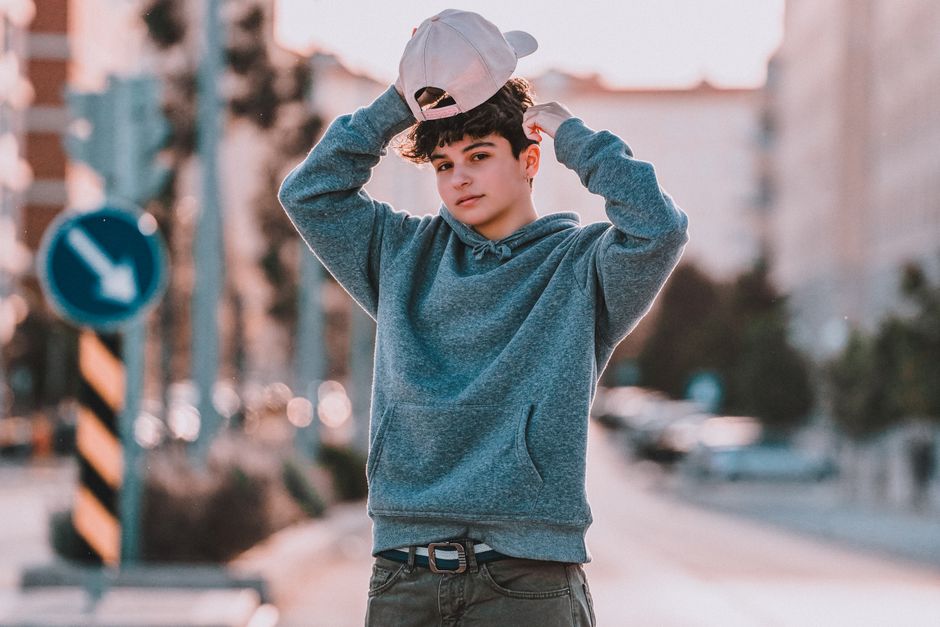 Young trendy male teenager in hoodie with cap standing with raised arms and looking at camera in town