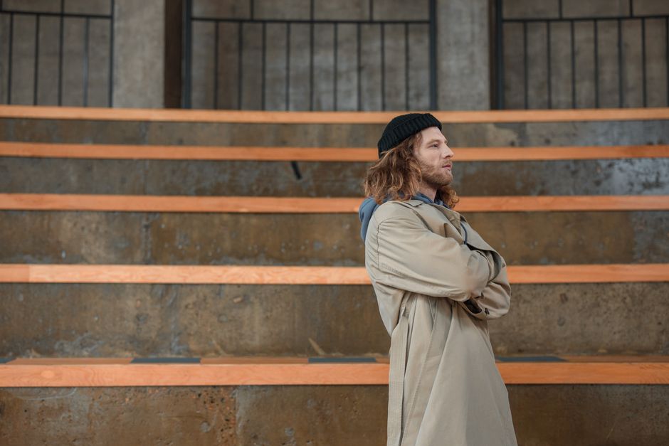 Man in a beige coat and beanie standing confidently outdoors on concrete bleachers.