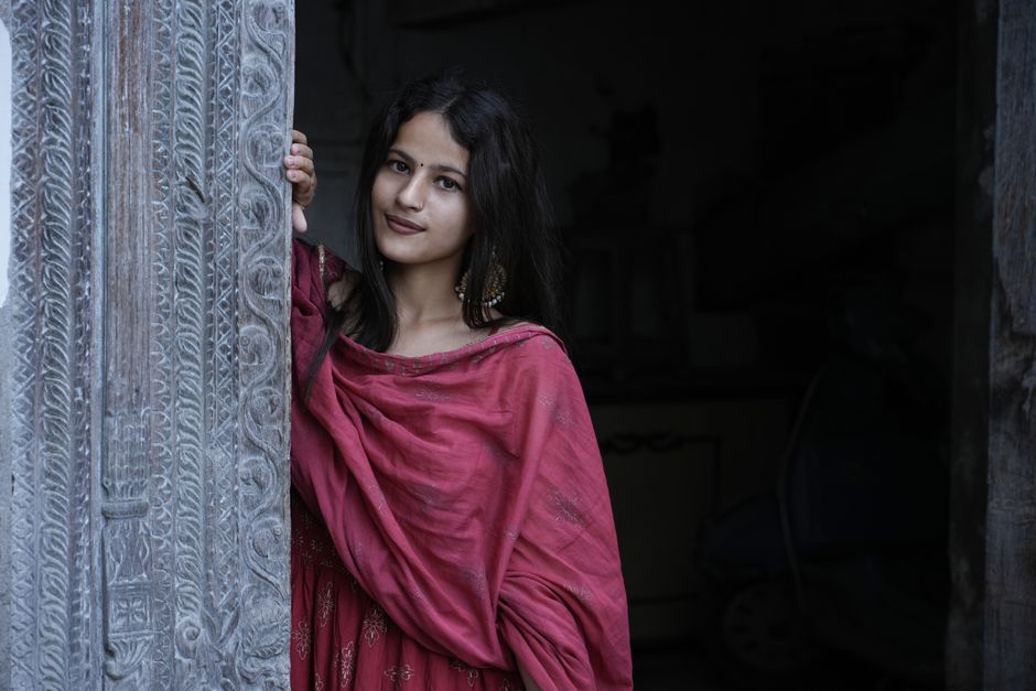 A woman in a vibrant traditional dress poses gracefully by a beautifully carved doorway.