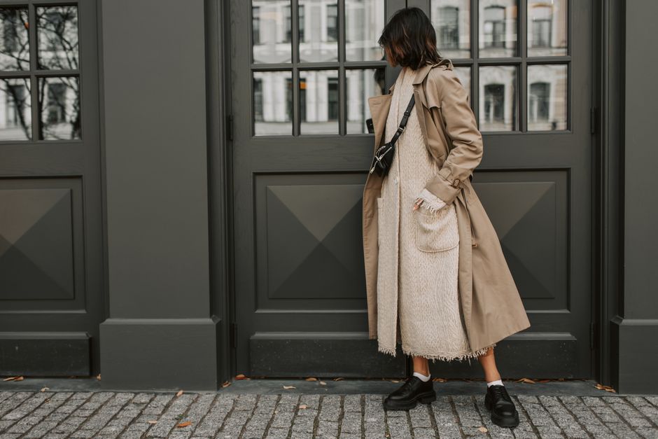 Woman in a trench coat leans casually against a modern urban background.