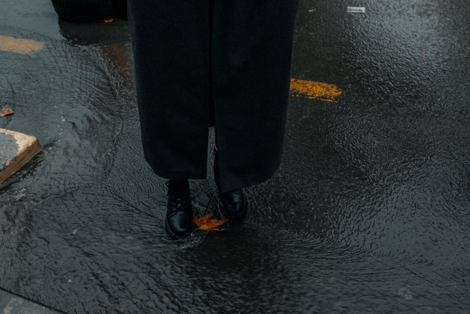 Person in black coat standing on wet city street during autumn rain.